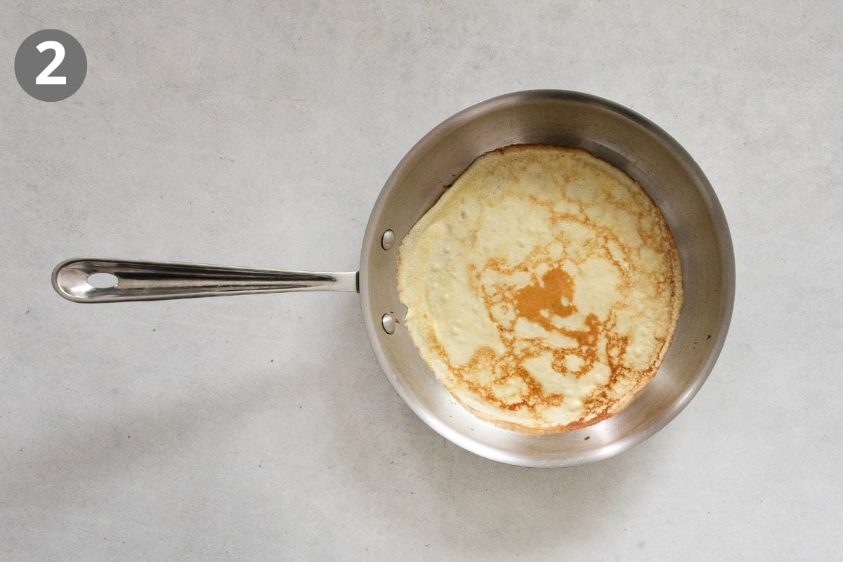 A thin swedish pancake cooking in a stainless steel skillet, shown from above on a light countertop. A small number 2 is in a gray circle in the upper left corner.