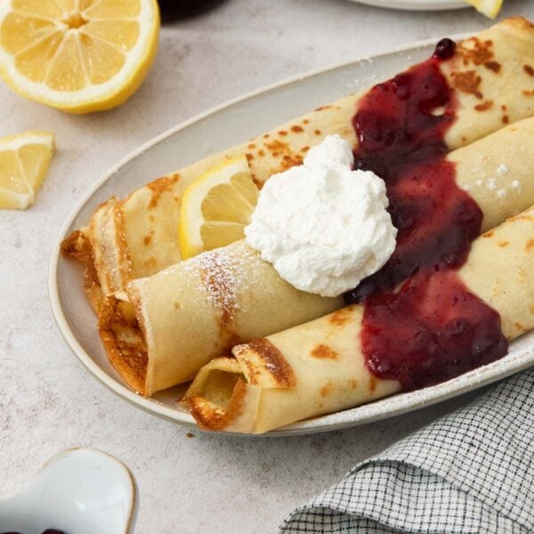 Three rolled swedish pancakes on a plate, topped with whipped cream, berry sauce, and a lemon slice. A halved lemon and napkin are nearby.