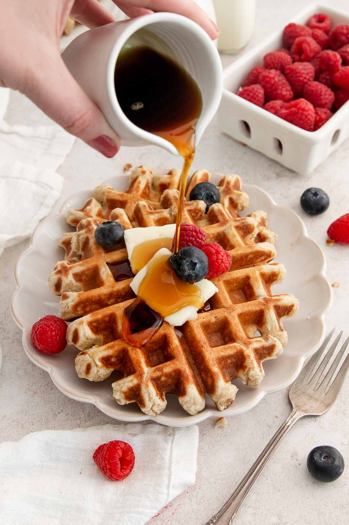 Hand pouring syrup onto waffles topped with butter, raspberries, and blueberries, with a fork and a carton of fresh raspberries nearby on a light table.