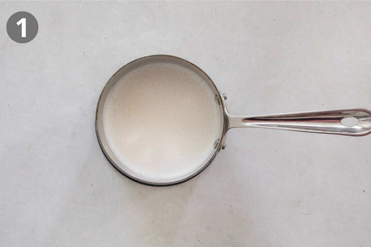 Small metal saucepan filled with milk, viewed from above, on a light-colored surface. A gray circle with the number 1 is in the upper left corner.