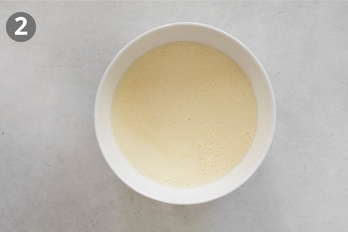 A white bowl filled with a pale, frothy liquid sits on a light gray surface. A gray circle with the number 2 is in the top left corner of the image.