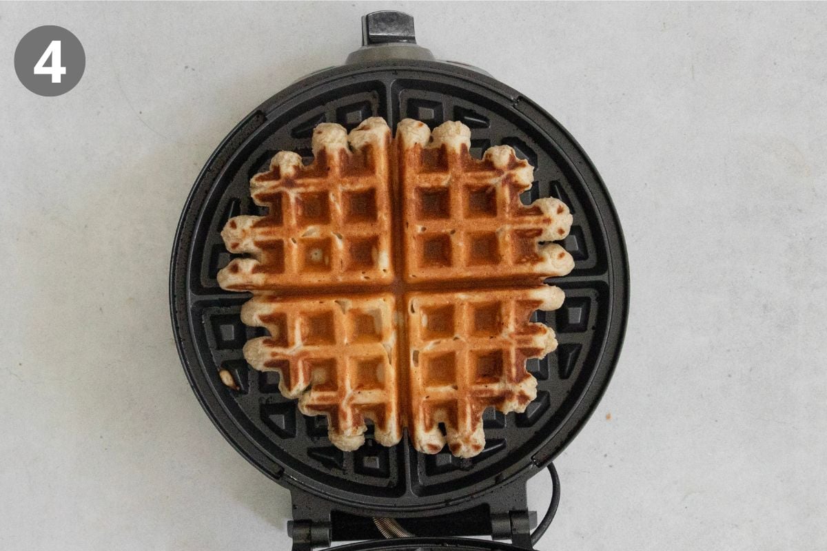 A golden brown waffle sits inside an open round waffle maker on a light surface. A gray circle with the number 4 is in the top left corner of the image.