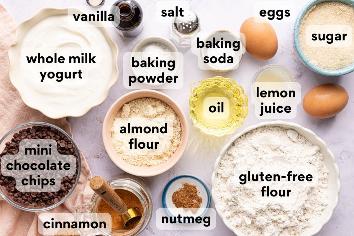 Various labeled baking ingredients are arranged on a white surface, including eggs, yogurt, flour, almond flour, oil, lemon juice, mini chocolate chips, cinnamon, nutmeg, baking powder, baking soda, salt, vanilla, and sugar.