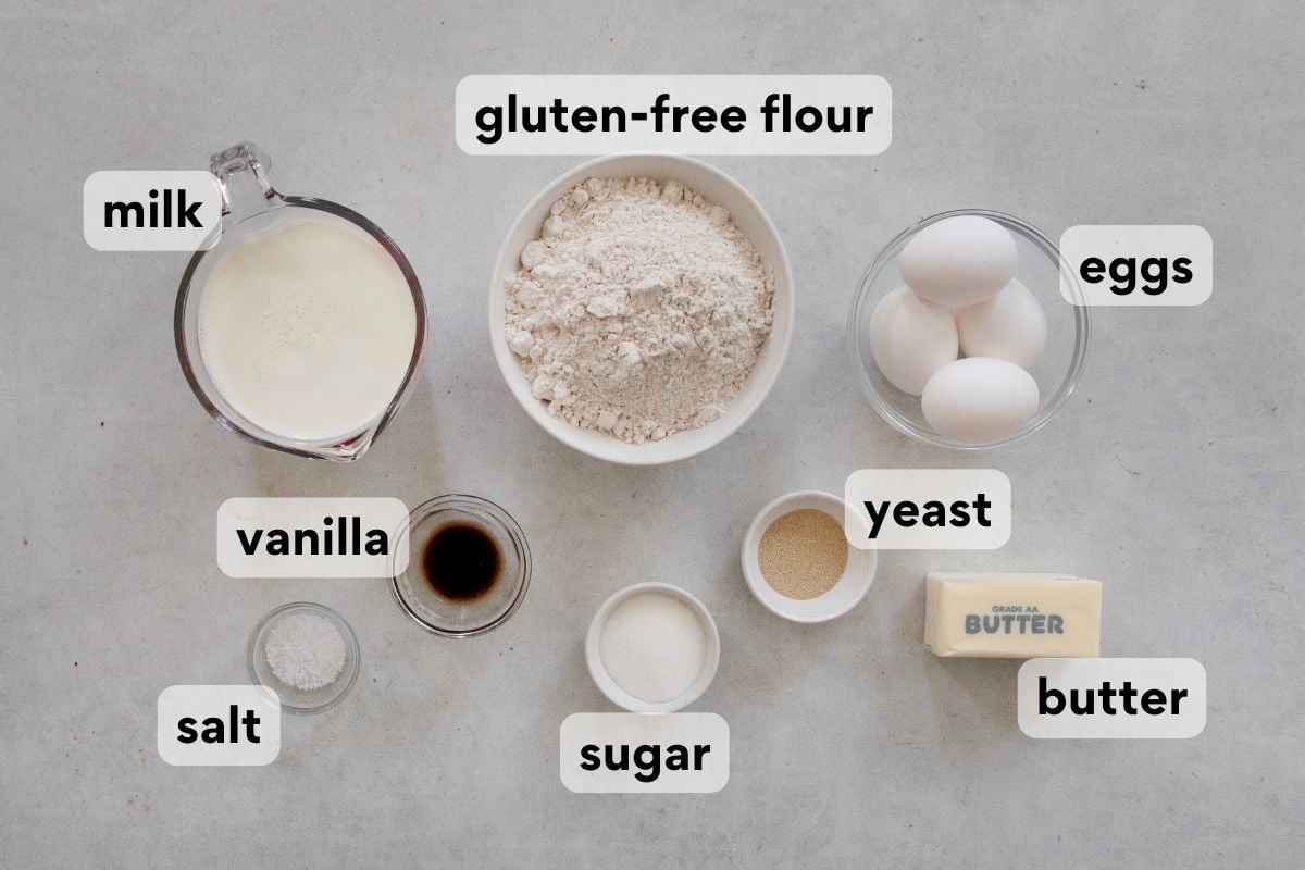 Measured ingredients for baking—milk, gluten-free flour, eggs, vanilla, yeast, butter, salt, and sugar—are arranged on a table. Each ingredient is labeled with white text on a black background.