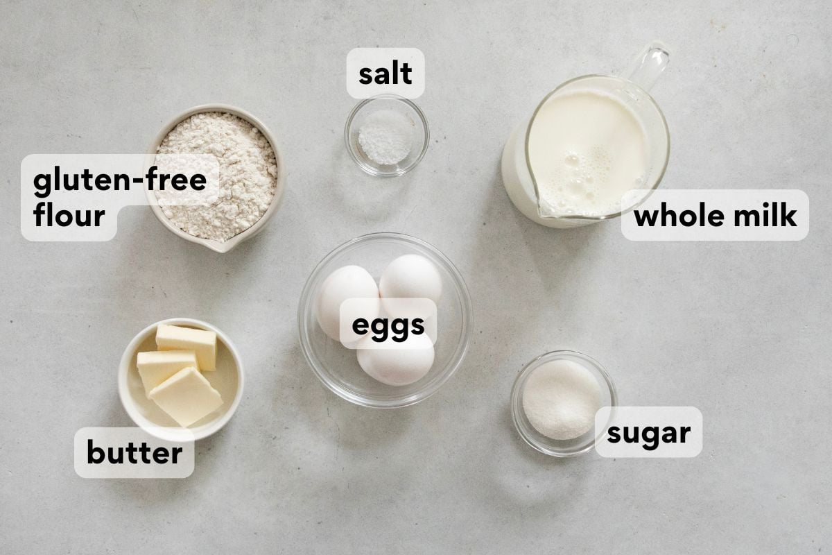 A flat lay of labeled baking ingredients: gluten-free flour, butter, eggs, sugar, salt, and whole milk arranged on a light surface.