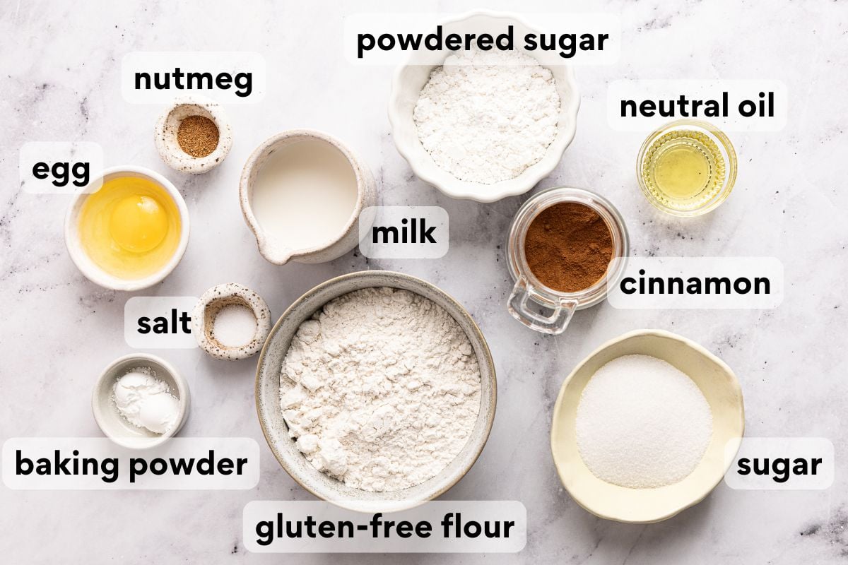 Top-down view of labeled baking ingredients: egg, nutmeg, salt, baking powder, gluten-free flour, milk, powdered sugar, neutral oil, cinnamon, and sugar, arranged on a white surface.