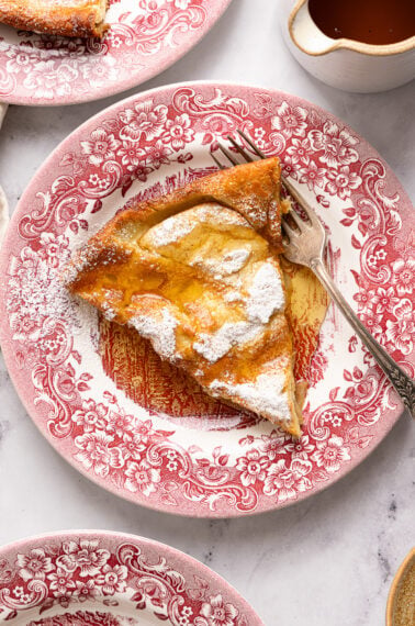 A slice of Dutch baby pancake topped with powdered sugar sits on a red floral plate, with a fork beside it and a small pitcher of syrup nearby.