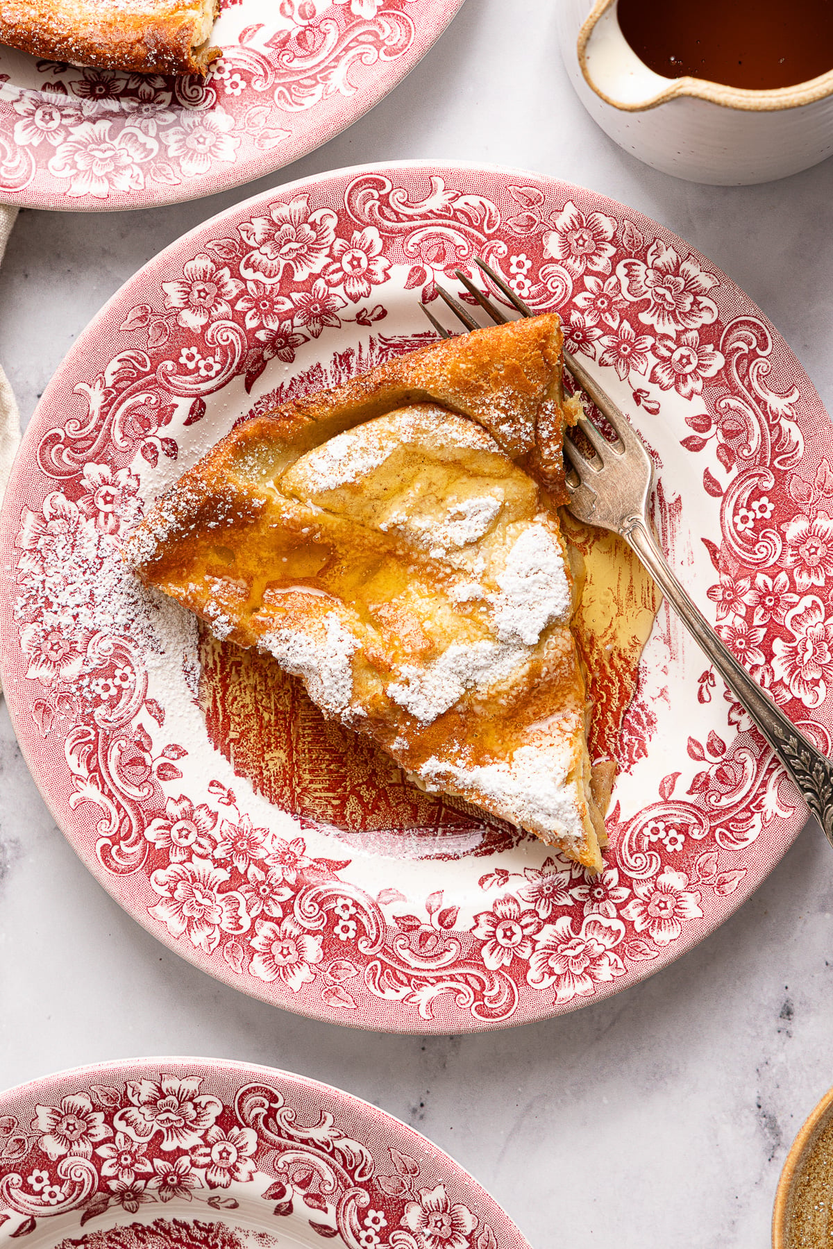 A slice of Dutch baby pancake topped with powdered sugar sits on a red floral plate, with a fork beside it and a small pitcher of syrup nearby.