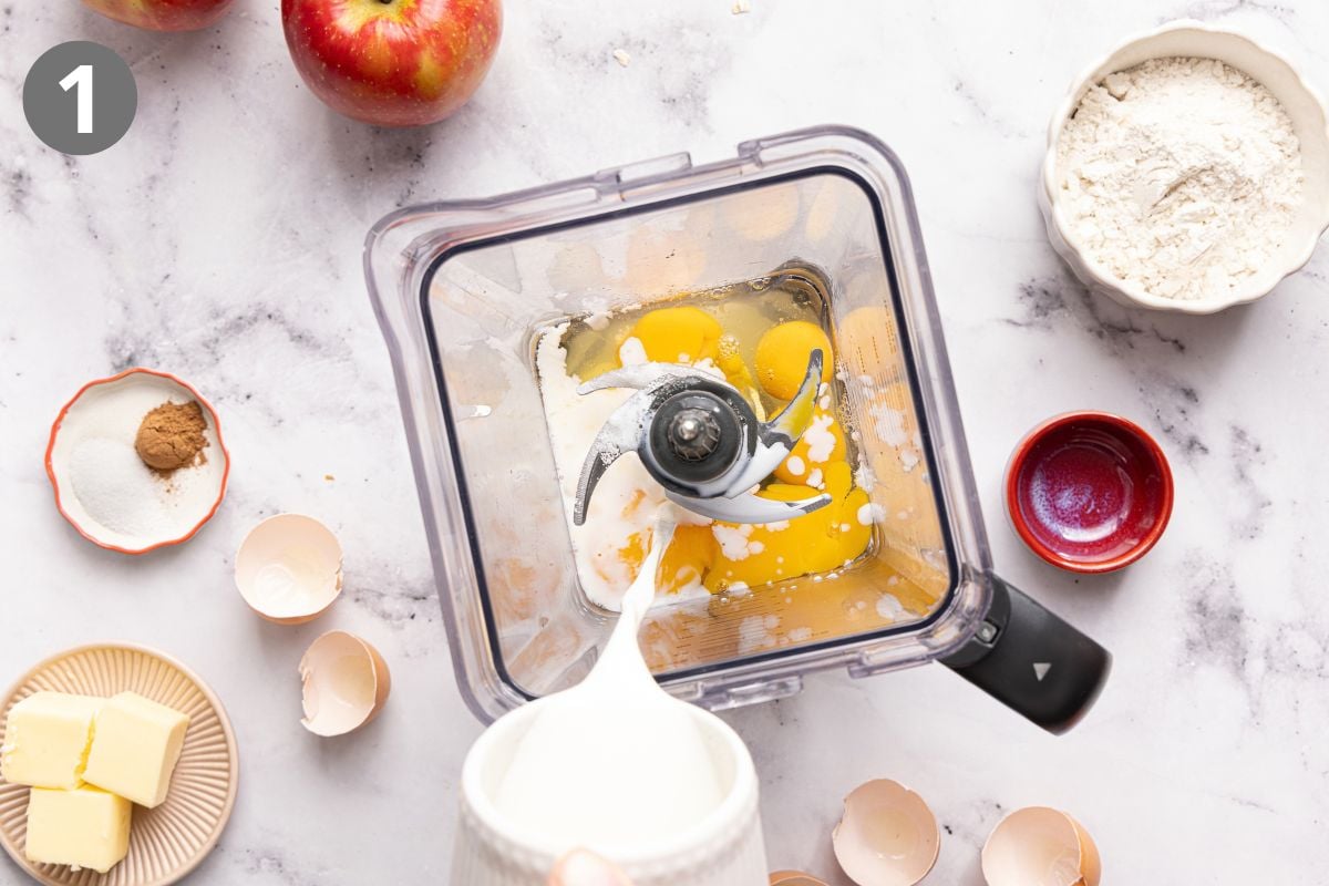 A blender filled with eggs and milk is seen from above, surrounded by apples, flour, butter, spices, sugar, egg shells, and a small red bowl, all on a marble countertop.