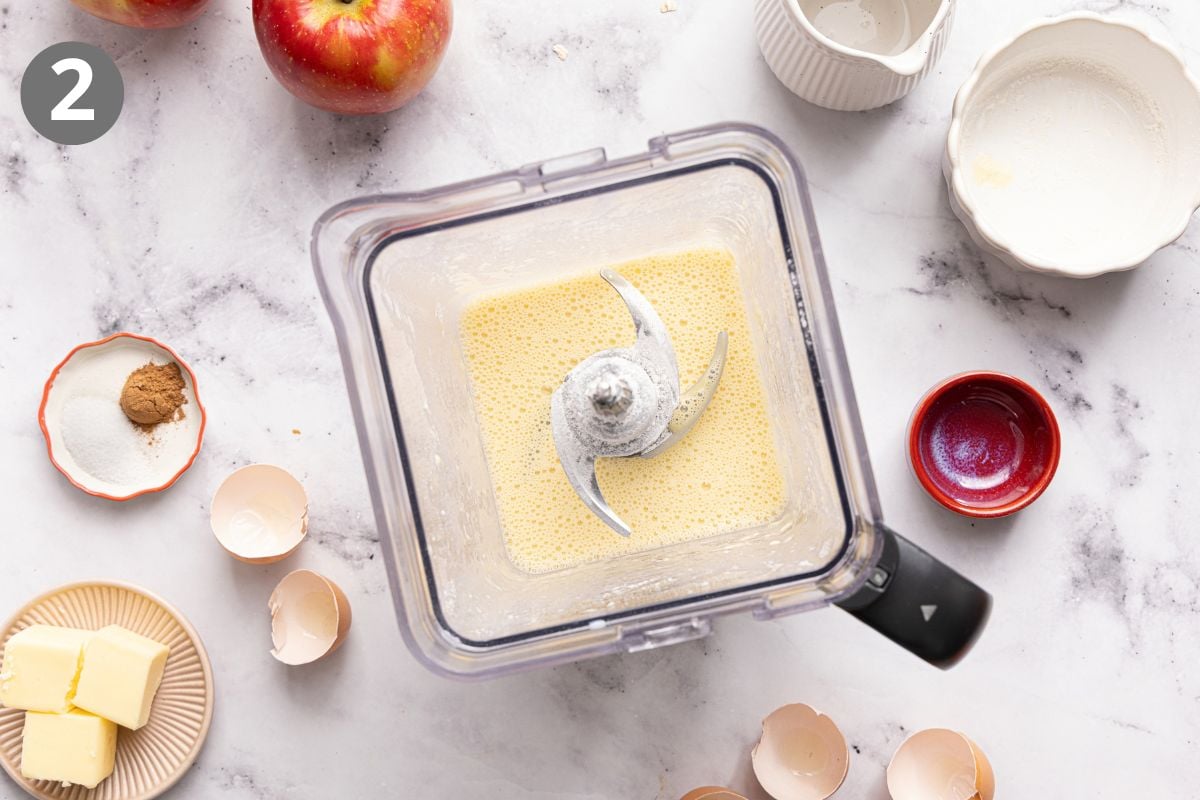 A blender with a creamy mixture is surrounded by empty bowls, cracked eggshells, butter, sugar, cinnamon, and apples on a marble surface. A gray circle with the number 2 is in the top left corner.