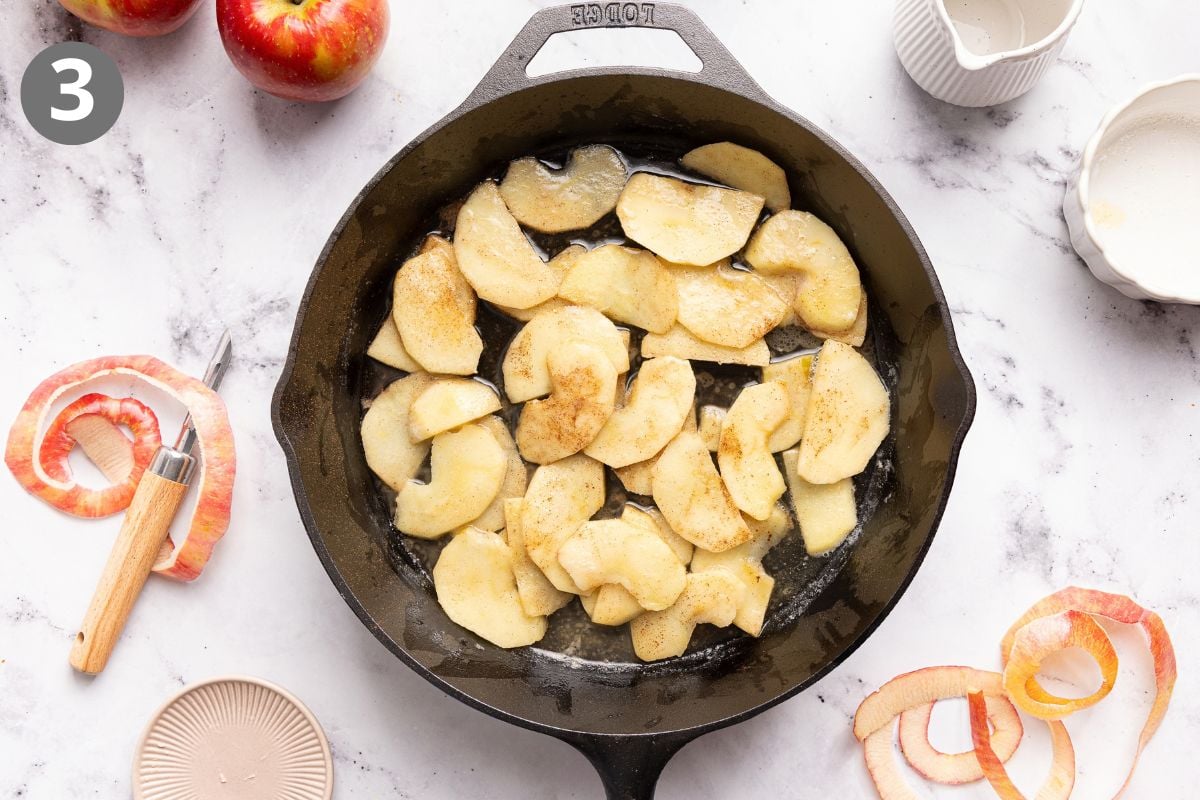 A cast iron skillet with sliced apples and cinnamon on a marble counter, surrounded by apple peels, a peeler, apples, and small dishes. A 3 is in the upper left corner.