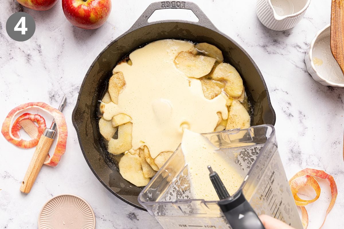 A blender pours batter over sliced apples in a cast iron skillet. Apple peels, whole apples, a peeler, and kitchenware are scattered on a marble countertop.
