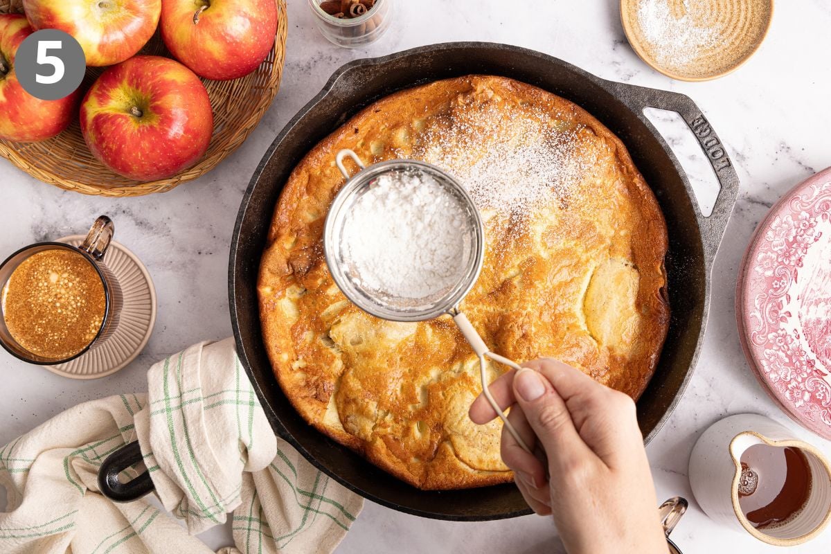 A hand sprinkles powdered sugar over a baked apple dutch baby pancake in a cast iron skillet. Nearby are fresh apples, a cup of coffee, a striped towel, a syrup pitcher, and a pink plate on a marble countertop.
