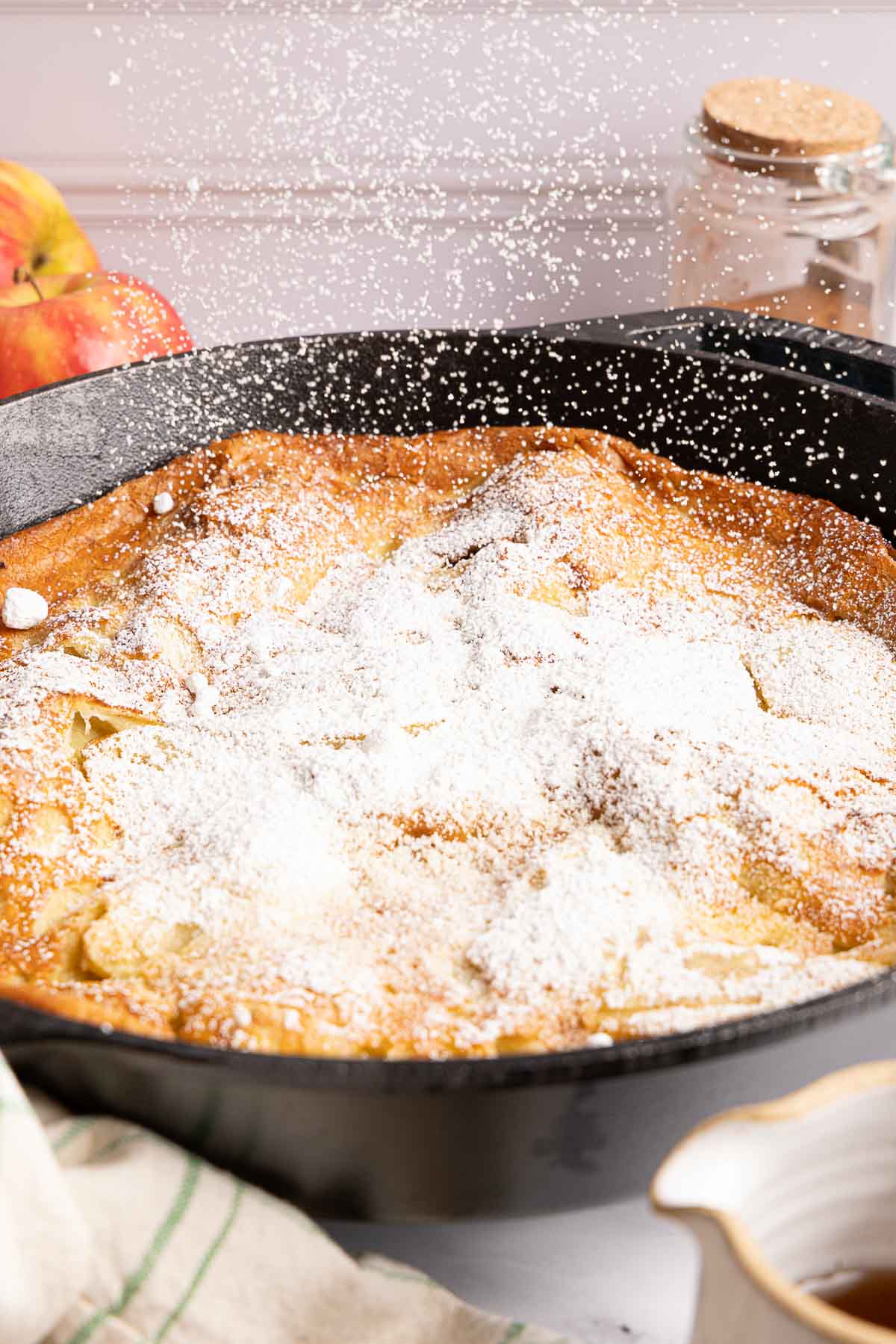 A golden-brown apple dutch baby in a black skillet is being dusted with powdered sugar, with apples and a jar in the background.