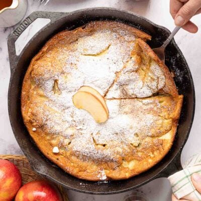 A skillet with a baked Dutch baby pancake, dusted with powdered sugar and topped with apple slices. A hand holds a spatula, ready to serve. Fresh apples are nearby on the table.