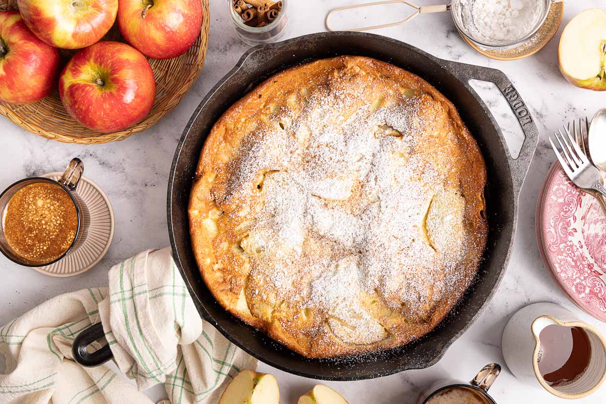 A baked apple dutch baby dusted with powdered sugar in a cast iron skillet, surrounded by apples, coffee, cinnamon sticks, plates, utensils, a napkin, and a syrup pitcher on a marble surface.
