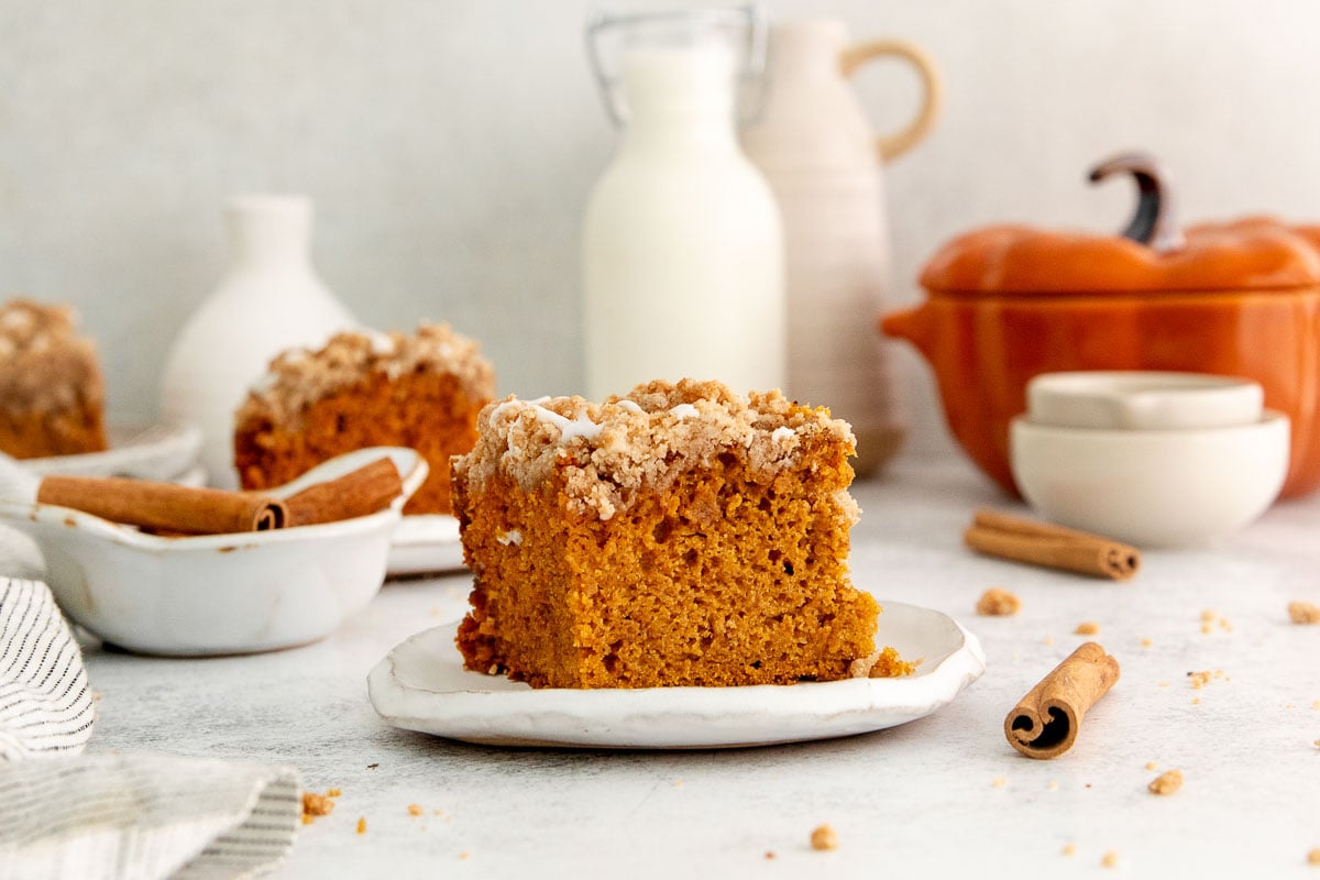 A slice of pumpkin crumb cake with crumb topping on a plate, surrounded by cinnamon sticks, a milk bottle, and autumn-themed dishes in a cozy setting.
