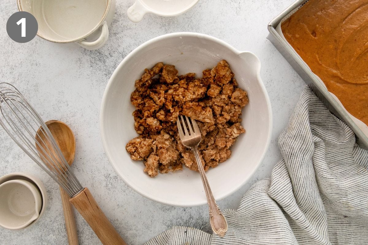 A white bowl with crumb topping mixture and a fork sits on a counter, surrounded by a whisk, wooden spoon, empty cups, a striped cloth, and a pan of batter.