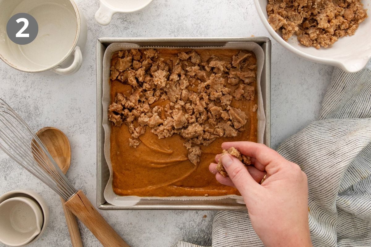 A hand sprinkles crumb topping over pumpkin batter in a square pan. Nearby are mixing bowls, a whisk, a wooden spoon, and a striped towel on a light countertop.