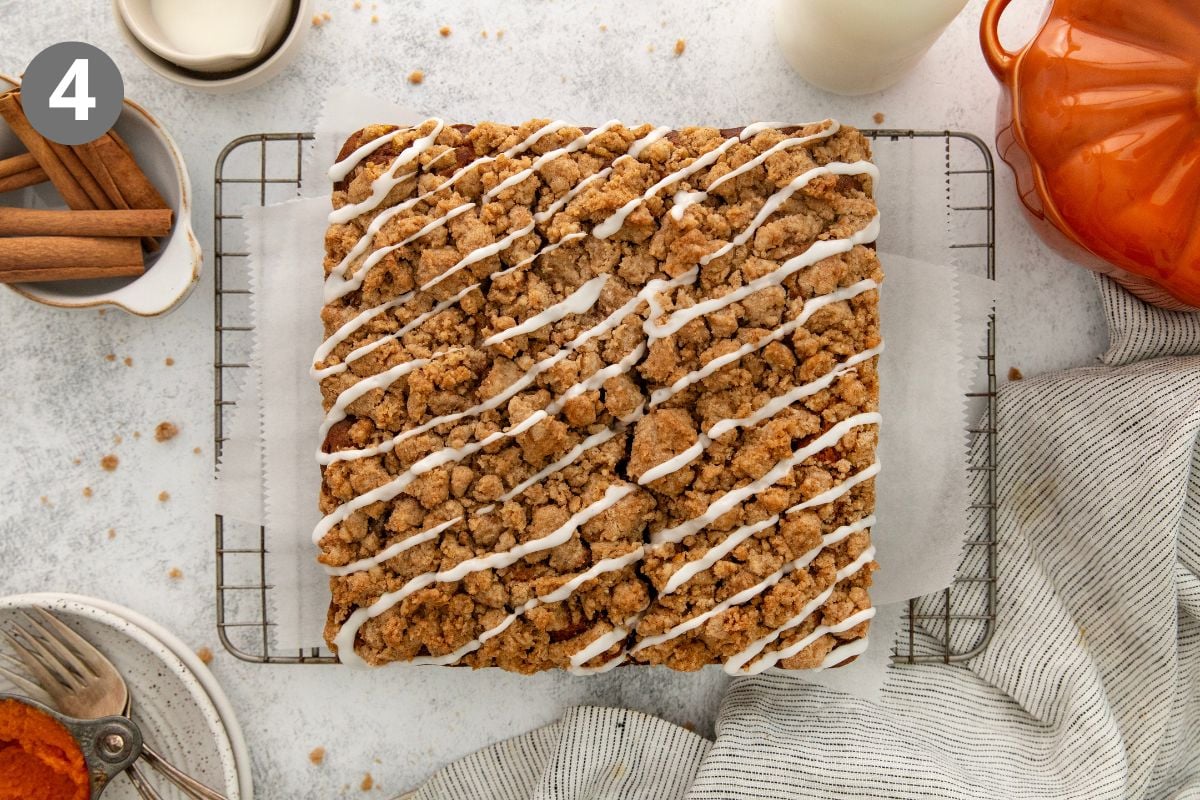 A large rectangular crumb cake with white icing drizzle sits on a wire rack lined with parchment paper, surrounded by a glass of milk, cinnamon sticks, and a striped napkin.