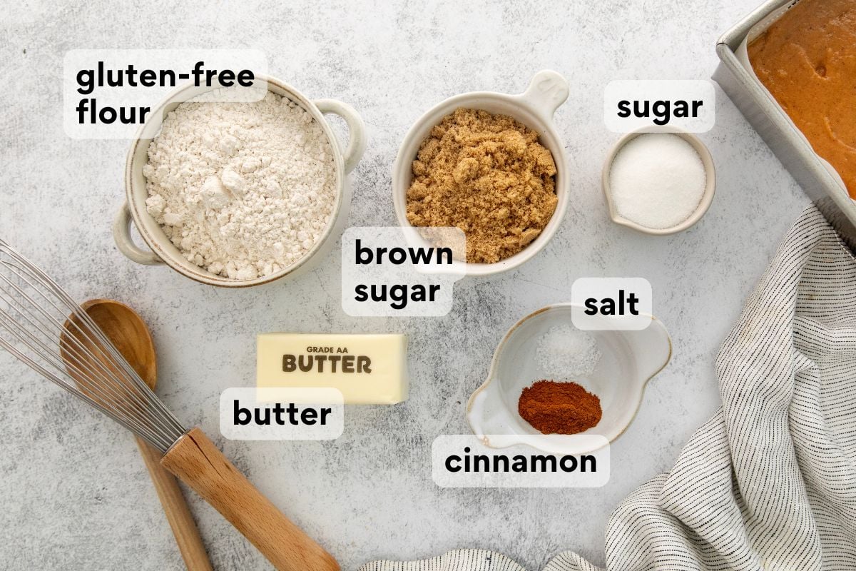Various baking ingredients on a table, including labeled bowls of gluten-free flour, brown sugar, sugar, salt, cinnamon, and a stick of butter, with a whisk and wooden spoon nearby.