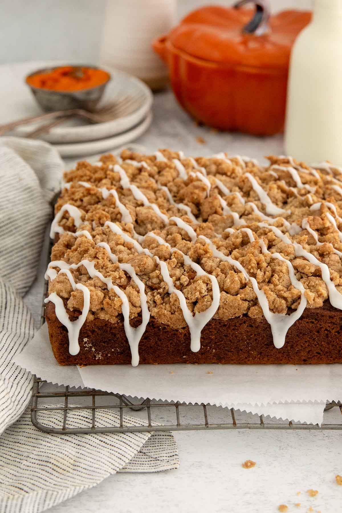 A rectangular pumpkin crumb cake sits on a cooling rack, topped with streusel and white icing drizzle. A striped towel, plates, orange dish, and milk bottle are in the background.