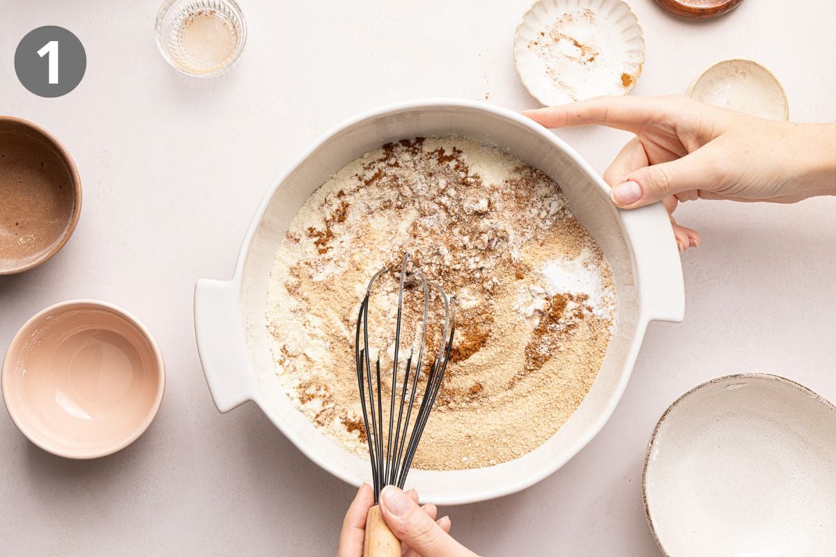 Hands holding a white mixing bowl with flour and brown sugar, using a whisk to combine the ingredients. Several empty bowls and dishes sit nearby on a light surface.