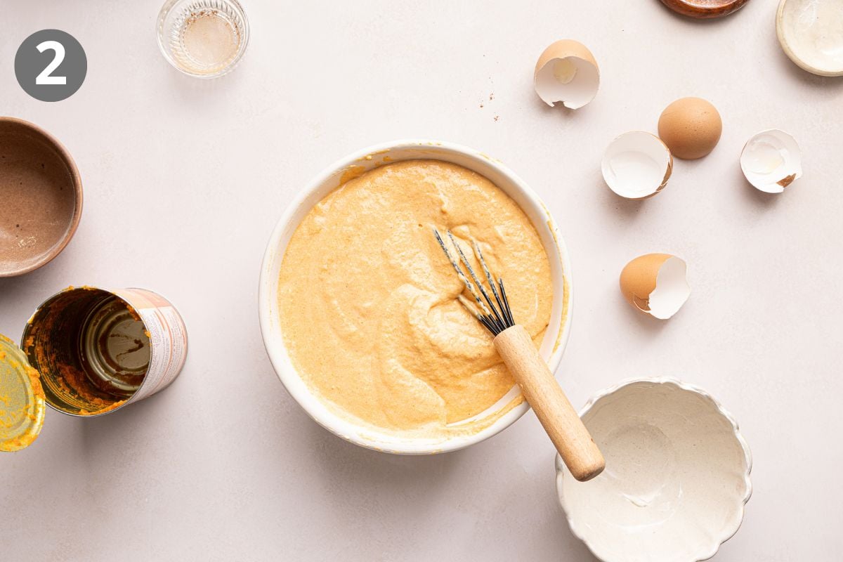 A mixing bowl with batter and a whisk, surrounded by eggshells, an empty can, small bowls, and a glass. The number 2 is in a gray circle in the top left corner.