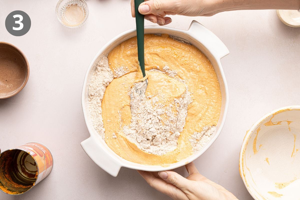 A person mixing flour into a yellow batter in a white bowl with a green spatula, step 3 in a recipe. Small bowls with ingredients and an open can are nearby on a light surface.