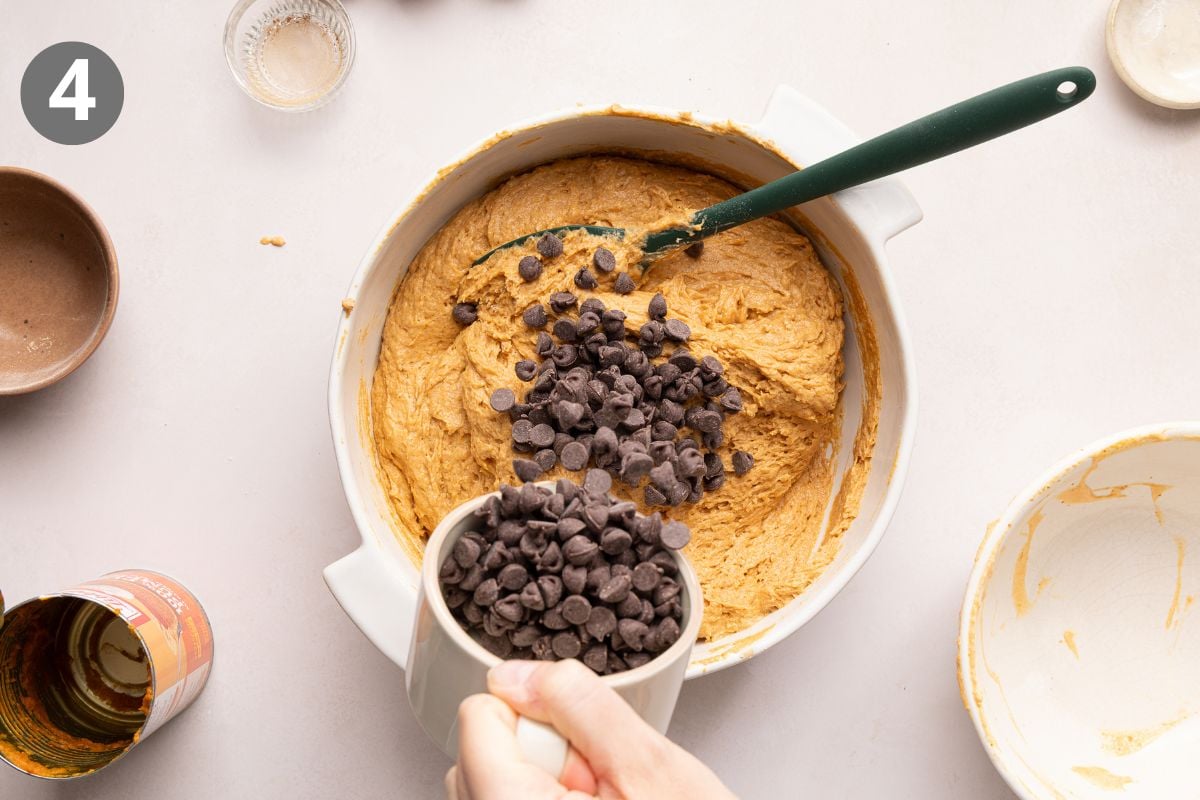 A hand pours chocolate chips from a white cup into pumpkin muffin batter in a mixing bowl with a green spatula. Several small bowls and an open can are nearby.