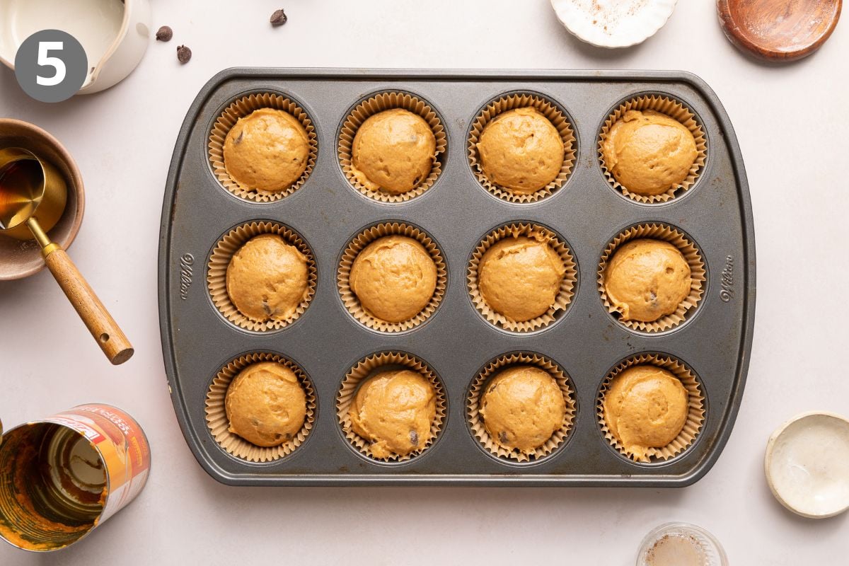 Pumpkin protein muffin batter in a non-stick muffin pan, surrounded by various baking utensils and empty bowls on a light countertop.