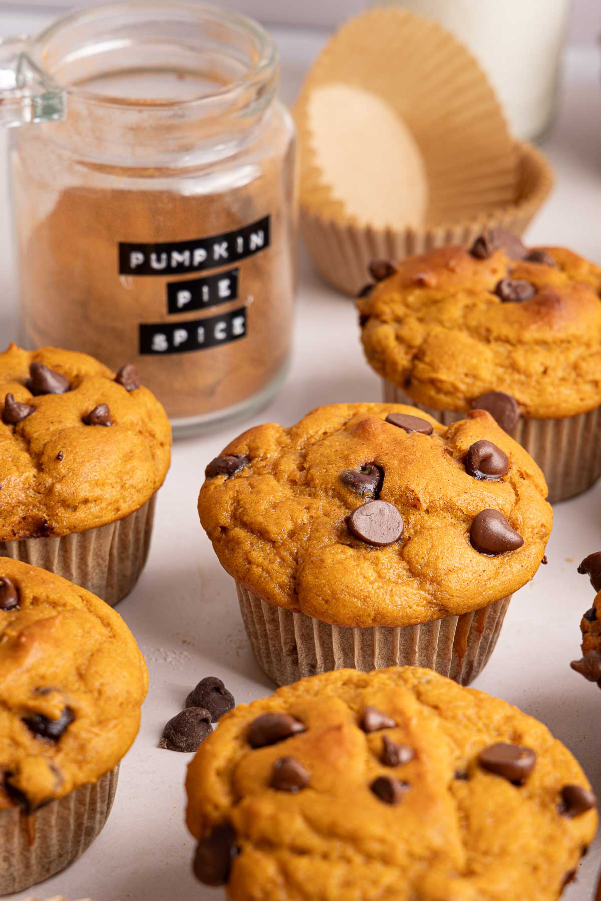 Pumpkin protein muffins on a white surface, with a glass jar labeled Pumpkin Pie Spice, an empty muffin liner, and a glass of milk in the background.