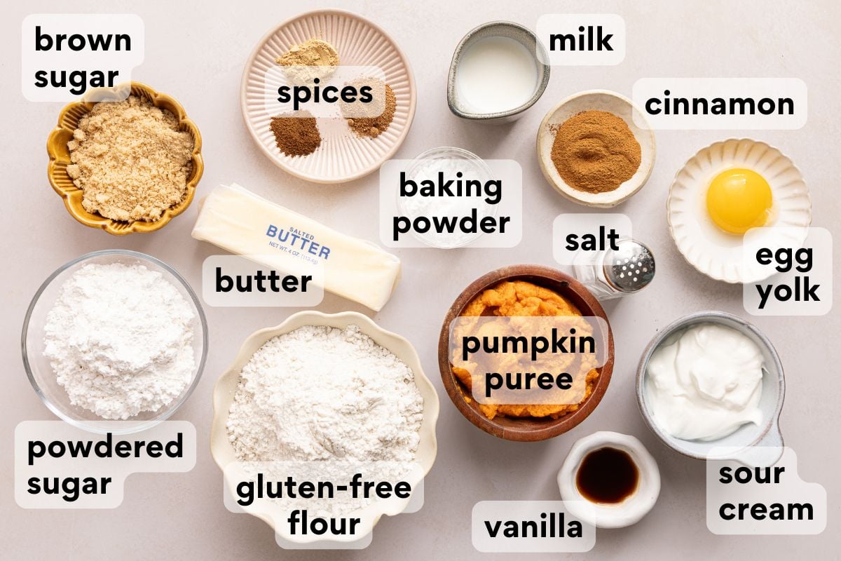 Labeled ingredients on a table for baking: brown sugar, spices, baking powder, butter, gluten-free flour, powdered sugar, milk, cinnamon, salt, egg yolk, pumpkin puree, sour cream, and vanilla.