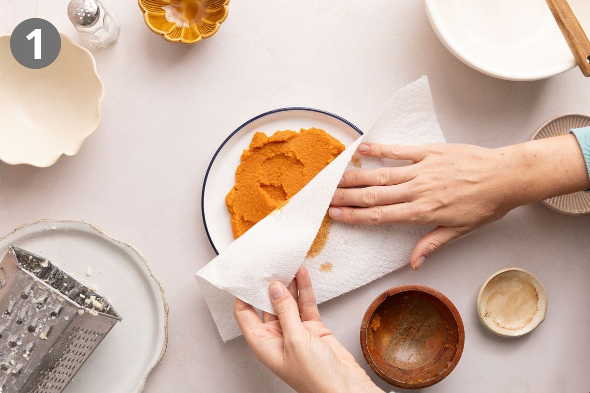 A person presses a paper towel onto a plate of pumpkin puree on a white table, surrounded by various empty bowls and a grater.
