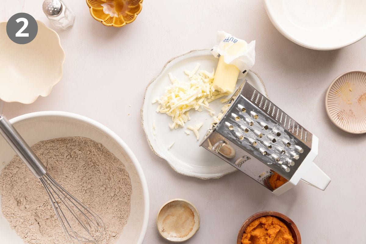 Overhead view of grated butter on a plate next to a box grater, with a bowl of flour mixture, a whisk, pumpkin puree, and various bowls on a white surface. The number 2 is in the upper left corner.