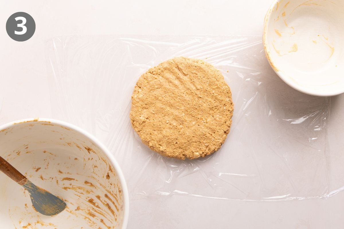 Round disc of dough on plastic wrap, surrounded by two empty mixing bowls—one with a spatula and some dough remnants. The number 3 is in the top left corner.