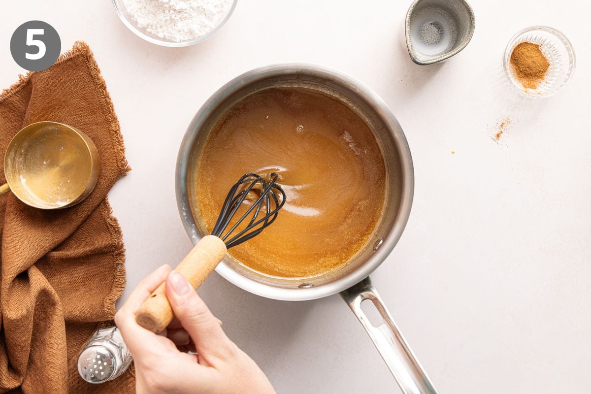 A hand whisks a brown sugar glaze in a saucepan. Nearby are a brown cloth, a small gold bowl, flour, spices, and a salt shaker. A circle with the number 5 is in the top left corner.