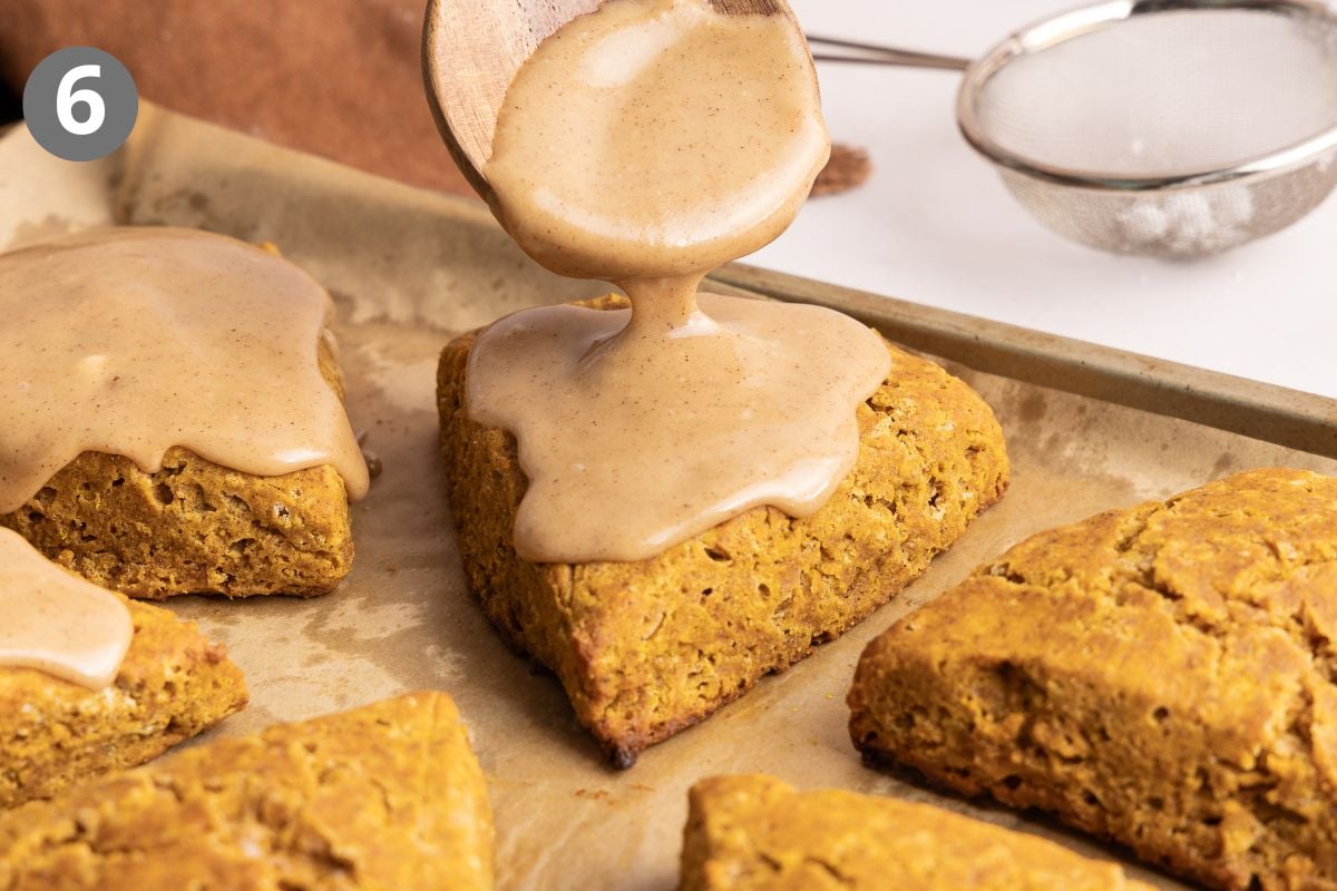 A wooden spoon drizzles brown sugar glaze over a triangular scone on a baking sheet, surrounded by other scones. A metal sieve is visible in the background.