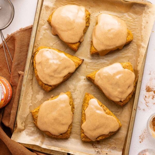 Six triangular scones with a tan glaze are arranged on a parchment-lined baking sheet. A brown cloth and kitchen tools are nearby, and the scene suggests a cozy, homemade baked good setting.