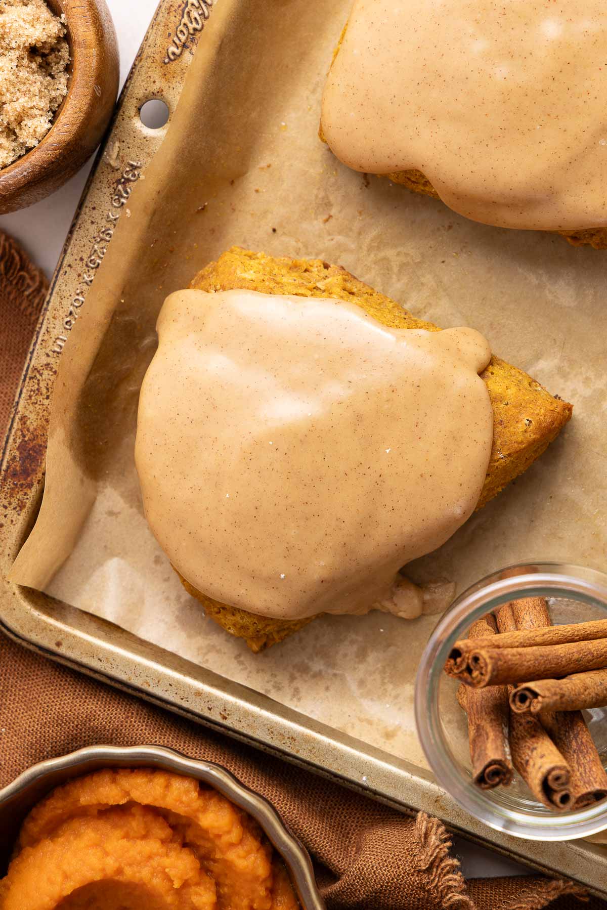 Pumpkin scone topped with a brown sugar glaze on a baking sheet, with cinnamon sticks, brown sugar, and pumpkin puree nearby.