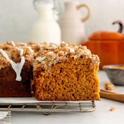 A close-up of a sliced pumpkin crumb cake with icing drizzle on a cooling rack. Milk and ceramic jugs are blurred in the background.