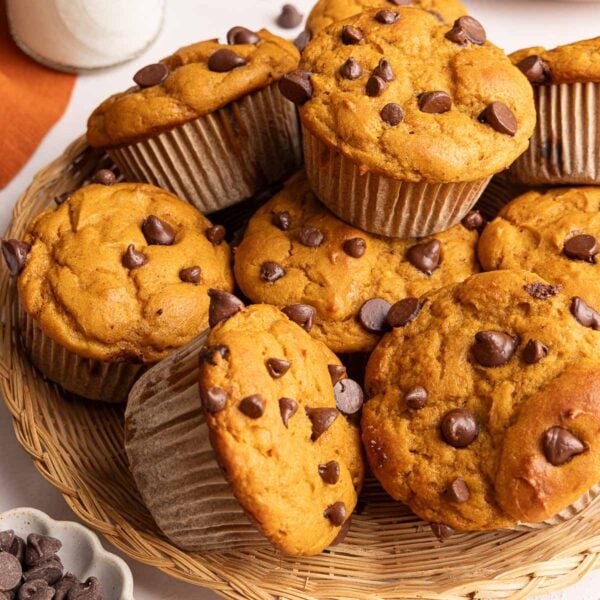 A wicker basket filled with pumpkin protein muffins topped with chocolate chips, with a small bowl of chocolate chips nearby.