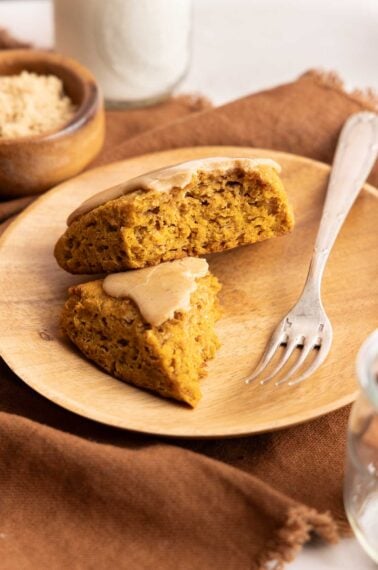 A wooden plate holds two triangular pieces of pumpkin scone with light brown glaze, next to a fork. A bowl of brown sugar and a glass of milk are in the background on a brown cloth.