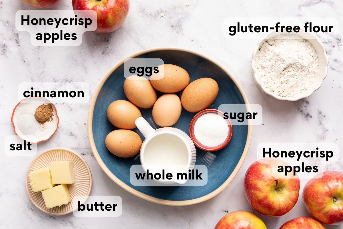 Top-down view of labeled baking ingredients: eggs, whole milk, sugar, gluten-free flour, butter, salt, cinnamon, and several Honeycrisp apples on a light surface. All items are arranged neatly.