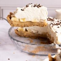 A slice of banoffee pie topped with whipped cream and chocolate shavings being lifted from a glass pie dish.