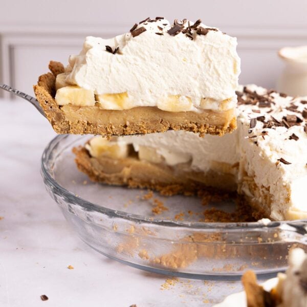 A slice of banoffee pie topped with whipped cream and chocolate shavings being lifted from a glass pie dish.