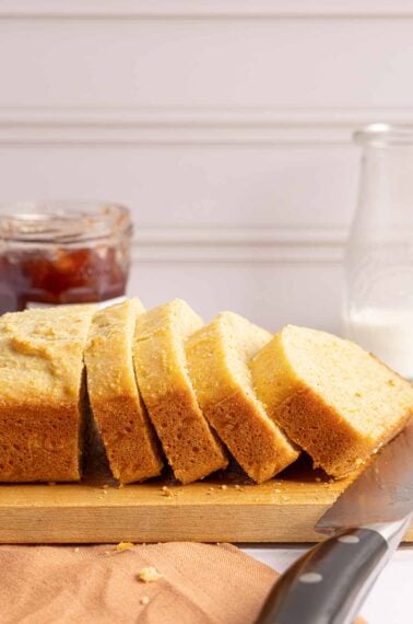 A sliced loaf of golden quick bread on a wooden cutting board, with a jar of jam and a glass of milk in the background.