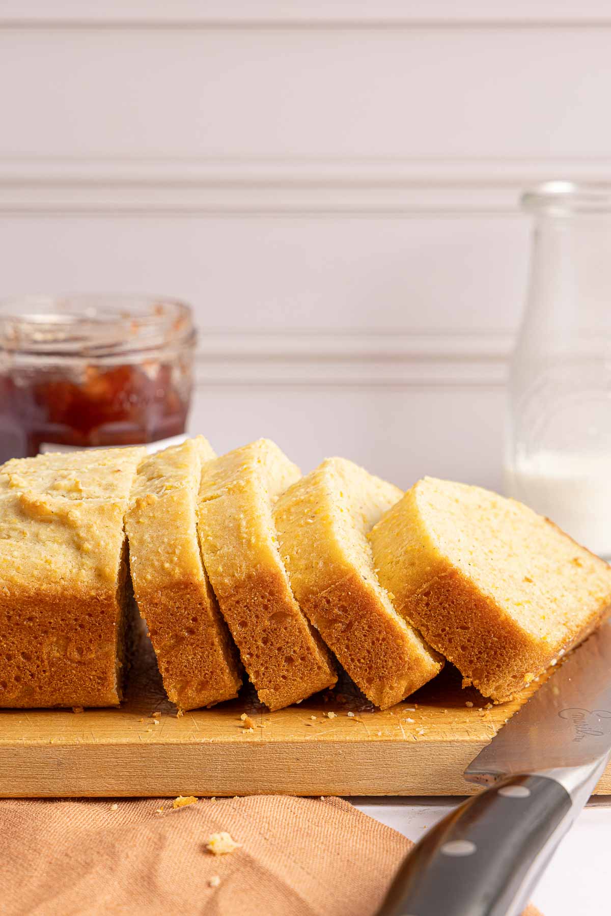 A sliced loaf of golden quick bread on a wooden cutting board, with a jar of jam and a glass of milk in the background.
