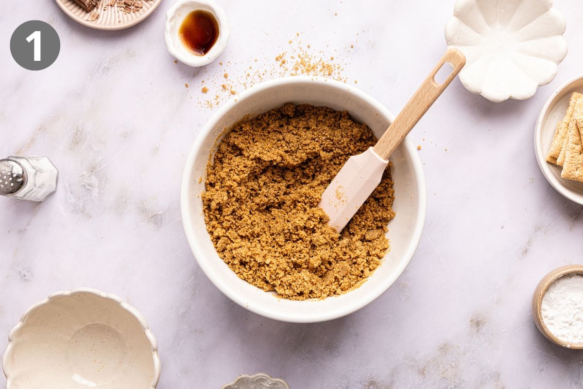 A white mixing bowl containing a mixture of graham cracker crumbs and butter with a spatula, surrounded by small dishes of ingredients.