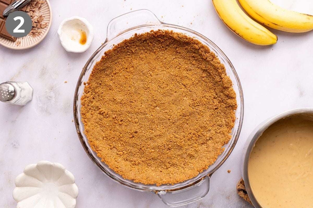 A graham cracker crust pressed into a clear glass pie dish, surrounded by a bowl of custard, bananas, and bowls with chocolate shavings and vanilla extract.